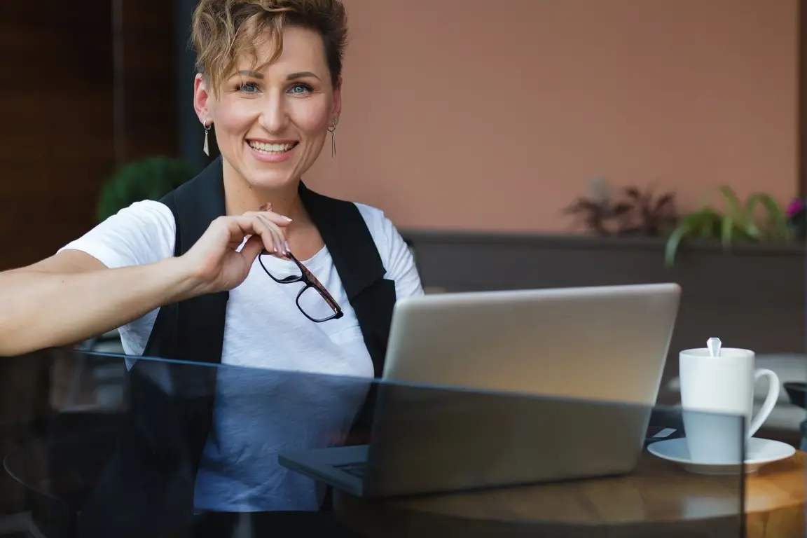 A woman sitting in front of her laptop, holding her glasses in her hand and smiling at the camera.