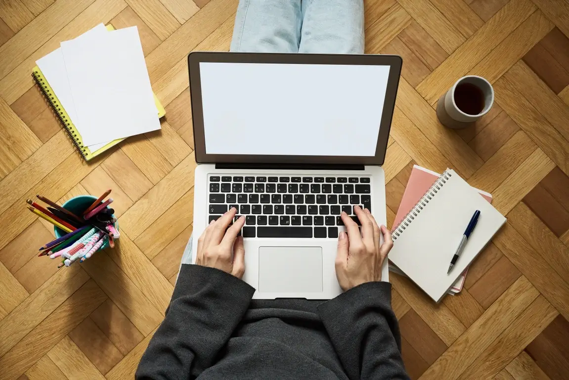 An overhead view of a laptop on someone's lap with pens and pencils, notebooks, and a mug of coffee on the floor around them.