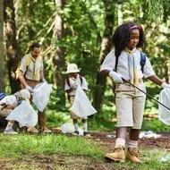 Young Black girl dressed in scouting uniform cleans up trash in the woods as part of community service with other den members.
