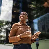 Black woman smiles up at sun, holding a coffee and her phone while walking outside.
