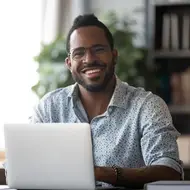 Black man smiles at a camera while working on his laptop at a desk