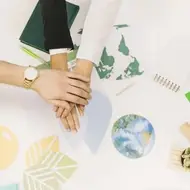 Imagem de 3 mãos unidas em cima de uma mesa com conteúdos sobre meio ambiente, cadernos e canetas.