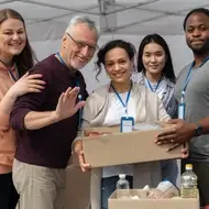 Foto de 5 pessoas voluntárias posando para a foto com a mulher ao centro segurando uma caixa com alimentos