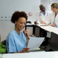 Black woman in blue scrubs sit behind desk at a health organization talking on the phone while looking at a report.