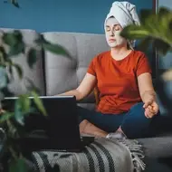 Woman sits on couch with hair up and face mask on practicing self care