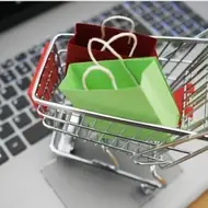 Imagem um mini carrinho de compra com duas sacolinhas feitas de dobradura de papel, uma verde limão e outra vermelha escura, em cima de um teclado de um notebook.
