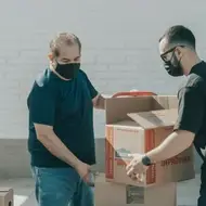 Imagem mostra 2 homens com mascara de proteção juntos ajudando no carregamento de caixas de papelão.