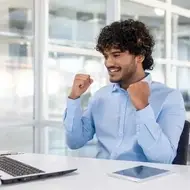 É uma foto de um rapaz jovem de cabelo encaracolado com camisa social, na frente de um notebook, com a cara contente parecendo que conseguiu ou conquistou algo.