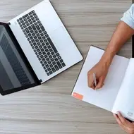 Hombre escribiendo en un cuaderno junto a su computadora