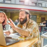 Um homem e uma mulher estão sentados juntos, conversando animadamente enquanto olham para um laptop em uma área de convivência iluminada.
