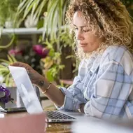 Black woman browses online continuing education courses at her desk at home with colorful plants in the background
