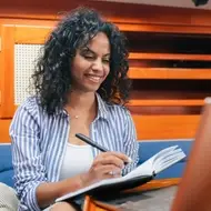 Woman writes in notebook in front of laptop, while sitting on a blue couch