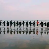 A group of 17 people holding hands in a long line at the beach