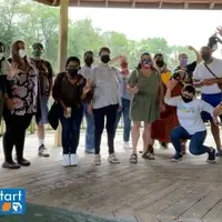 A group of multiracial people outdoors in a gazebo wearing face masks. Some are waving and some are making silly faces or gestures/poses. Fresh Start's logo is in the bottom left corner.