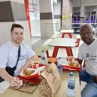 an Thomson, a law and human rights intern from Gonzaga University, Spokane, and Joseph Odoteye from Pro-Passion Projects sharing lunch at Accra Mall KFC during Ian’s orientation in Accra, Ghana. Both are wearing Pro-Passion Projects t-shirts. They are enjoying a meal of jollof rice and grilled chicken while discussing the internship program, orientation schedule, and upcoming community projects. The background shows the restaurant interior with other patrons and tables. This image captures a casual, friendly moment that highlights the supportive and welcoming environment of Pro-Passion Projects, emphasizing mentorship, relationship-building, and preparation for interns before they begin their volunteer and community engagement activities in Ghana."