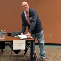 Bald man standing next to a table with paperwork in his hands.