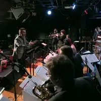 A man claps his hands in front of a group of students with musical instruments, while an audience listens.