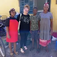 Shayla, a social work volunteer from the USA, pictured with a school-age boy who has dropped out of school, his mother, and staff from the La Community Social Welfare Department during an outreach program in La, Accra, Ghana. Shayla is standing alongside the team of social welfare officers from the La Municipal Assembly, who are identifying children of school-going age staying at home due to financial or social constraints. The mother appears attentive while the boy looks curious. The background shows a typical residential area in La with local homes and streets. This image captures Shayla actively engaging with the community, observing and assisting the social welfare team, and contributing to efforts aimed at improving school attendance and child welfare in the area.