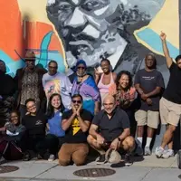 The image shows a group photo of the Diverse City Fund Grantmaking Team for 2023. The photograph was taken by Kayla Daley, as mentioned in the image file name.  The picture features a diverse group of individuals standing together in front of a colorful mural. The mural includes a large portrait of what appears to be Frederick Douglass, an important historical figure in American civil rights. The background of the mural has vibrant colors including orange, blue, and yellow.  The group consists of approximately 15-20 people of various ages, genders, and ethnicities, body ability reflecting the diversity mentioned in the organization's name. They are dressed casually, with some wearing t-shirts and others in more formal attire. The group members are smiling and appear to be in good spirits, conveying a sense of camaraderie and shared purpose.  This image represents the team responsible for making grant decisions for the Diverse City Fund.