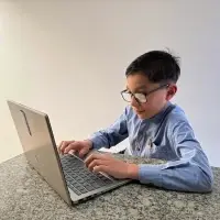 young boy from Colombia sitting at desk with laptop engaged in his online English class, smiling and wearing glasses