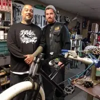 Two men pose with a chopper bicycle they are fixing.
