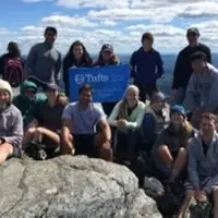A group of people pose with a Tufts banner sitting on rocks with an expansive vista behind them.