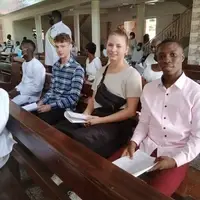 Stephen, Human Rights Coordinator with Pro-Passion Projects, pictured sitting inside a Catholic church in Ghana with two volunteers participating in childcare and veterinary projects. None of them are wearing Pro-Passion Projects t-shirts; all are dressed casually. The volunteers are seated attentively, discussing their upcoming activities, challenges, and experiences in the community. The church interior, with wooden pews and stained glass windows, provides a calm and reflective environment. This image captures a quiet moment of mentorship and planning, highlighting how Pro-Passion Projects supports volunteers’ understanding of local culture, community needs, and the holistic context of their childcare and veterinary work in Ghana