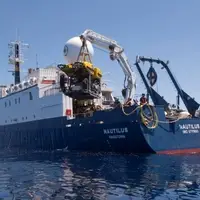 Exploration Vessel Nautilus viewed from the port quarter painted blue near the waterline and white above decks is launching a yellow Remotely Operated Vehicle using a white crane