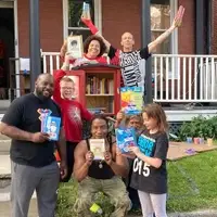A group of adults and children hold books and raise their arms in celebration around a red Little Free Library book-sharing box.