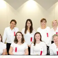 Eight women in white shirts and black pants behind a long table. The first row of four women are seated and the second row of women are standing behind them.