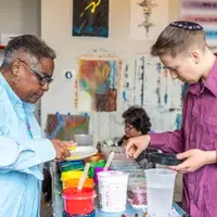 A Black woman in her 50's has gold hoops, short grey and black hair, and black glasses. She is wearing an oversized light blue button down shirt, and is using a popscile stick to scoop yellow paint onto a white plate. She stands in front of a narrow table of many colored paints in round plastic containers. Across from her is a white nonbinary person in their 20's with blonde hair and a yarlkmulke. They are wearing a maroon button down shirt, and are holding a black plastic container. Their hand is in midair as they decide what color to choose. The background shows a wall with paint splatters and a colorful artwork, as well as one woman sitting in the background.