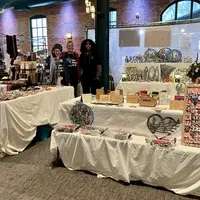 Three people stand in the corner behind three tables filled with Haitian art: metal wall art, bracelets, earrings, greeting cards, ceramic mugs, etc.