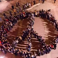 picture of a group of people creating a peace sign in a courtyard while holding their hands up. The picture is on a brown floored courtyard and there are about 300 people in the formation. The photo was taken from a bird's eye view.