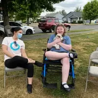 Heather is smiling at the camera and holding a sloth stuffed animal. A support staff member sits next to her. They are in a park.