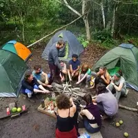 A group of international volunteers preparing breakfast over a campfire at a jungle campsite in the Peruvian Amazon. Tents are pitched under the rainforest canopy near Codo del Pozuzo, showing the immersive community life and outdoor adventure aspect of the Semillas Life NGO program.
