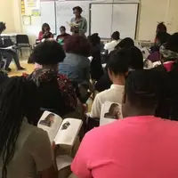 a teen author reads her book to a class full of students. The students have copies of the. book and are looking at the page.