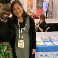 Two women stand in front of a table with brochures in a booth at a conference.