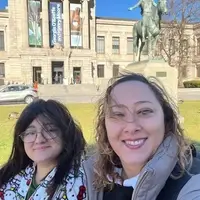 A mentee, pictured with long black hair and glasses, wearing a Hello Kitty zip up hoodie, and her mentor, pictured with long hair and wearing a silver coat, smiling at the camera, posing in front of the Museum of Fine Arts in Boston, MA.