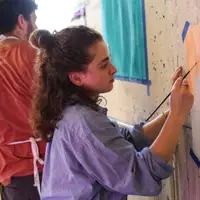 A young white woman i her twenties with curly brown hair wearing oversized blue button up shirt as she stands at a paint-speckled wall. She is holding up a paintbrush and making marks with orange paint on a white page