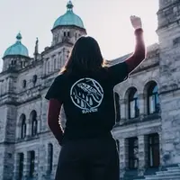 A person wearing a RAVEN branded t-shirt stands in front of the BC parliament buildings with their fist raised in the air