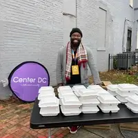 A smiling Black American Muslim man standing behind a table, distributing food on the corner in prepackaged boxes