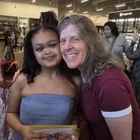 A girl wearing a light blue dress is standing with her mentor, a woman who is wearing a maroon shirt. They are both smiling and leaning into each other.