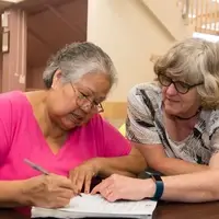 A woman writes in a workbook while a tutor leans in to help, in the Literacy Center.
