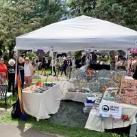 4 people stand to the left of a 10foot white canopy. A Progressive Pride flag is taped on the left front leg of the canopy. Three tables under the canopy display Haitian metal wall art, ceramic mugs, bracelets, and earrings.