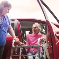 A volunteer driver helps a senior client using a walker step out of a vehicle. The volunteer provides support by holding the client’s arm, demonstrating safe and caring assistance.