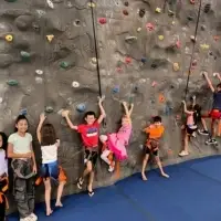 A group of elementary and middle-school aged children smile and play at the base of an indoor climbing wall.