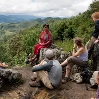 Guatemalan woman sits on a rock overlooking a valley, talking to 4 western students who are sat listening