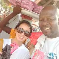 Victoria Ngo-Lam, a human rights volunteer from San Francisco, pictured with Joseph Odoteye from Pro-Passion Projects during her orientation at Makola Market in Accra, Ghana. Both are wearing Pro-Passion Projects t-shirts and enjoying yogurt under the hot afternoon sun while discussing her upcoming volunteer activities, orientation schedule, and community engagement projects. The bustling market environment is visible in the background, with vendors, colorful goods, and local shoppers adding context to the setting. This image captures a relaxed and friendly moment, highlighting Pro-Passion Projects’ supportive and inclusive orientation process that provides mentorship, guidance, and cultural immersion before hands-on volunteer work begins.