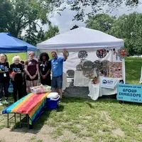 5 people stand to the left of a 10-foot white canopy. Two benches covered by a rainbow beach towel are in front of of the people, with a bin of "your choices" of free Fair Trade tea in boxes. Under the canopy: 3 tables full of Haitian art: metal wall art, ceramic mugs and vases, earrings, and bracelets.