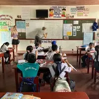 Two international volunteers at Semillas Life NGO teaching an interactive English lesson to a group of local primary school children in Codo del Pozuzo, Peruvian Amazon. The students are smiling and engaged with educational flashcards on a chalkboard.